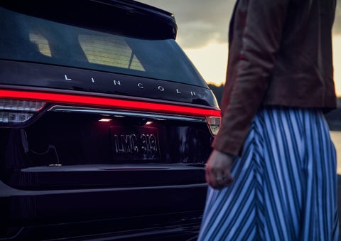 A person is shown near the rear of a 2024 Lincoln Aviator® SUV as the Lincoln Embrace illuminates the rear lights | Carman Lincoln in New Castle DE