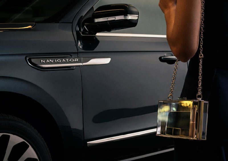 A woman is approaching a 2024 Lincoln Navigator® SUV with an elegant clear purse displaying a smartphone and lipstick.