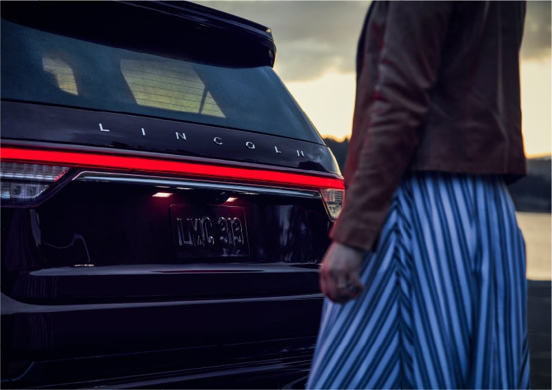 A person is shown near the rear of a 2023 Lincoln Aviator® SUV as the Lincoln Embrace illuminates the rear lights | Carman Lincoln in New Castle DE